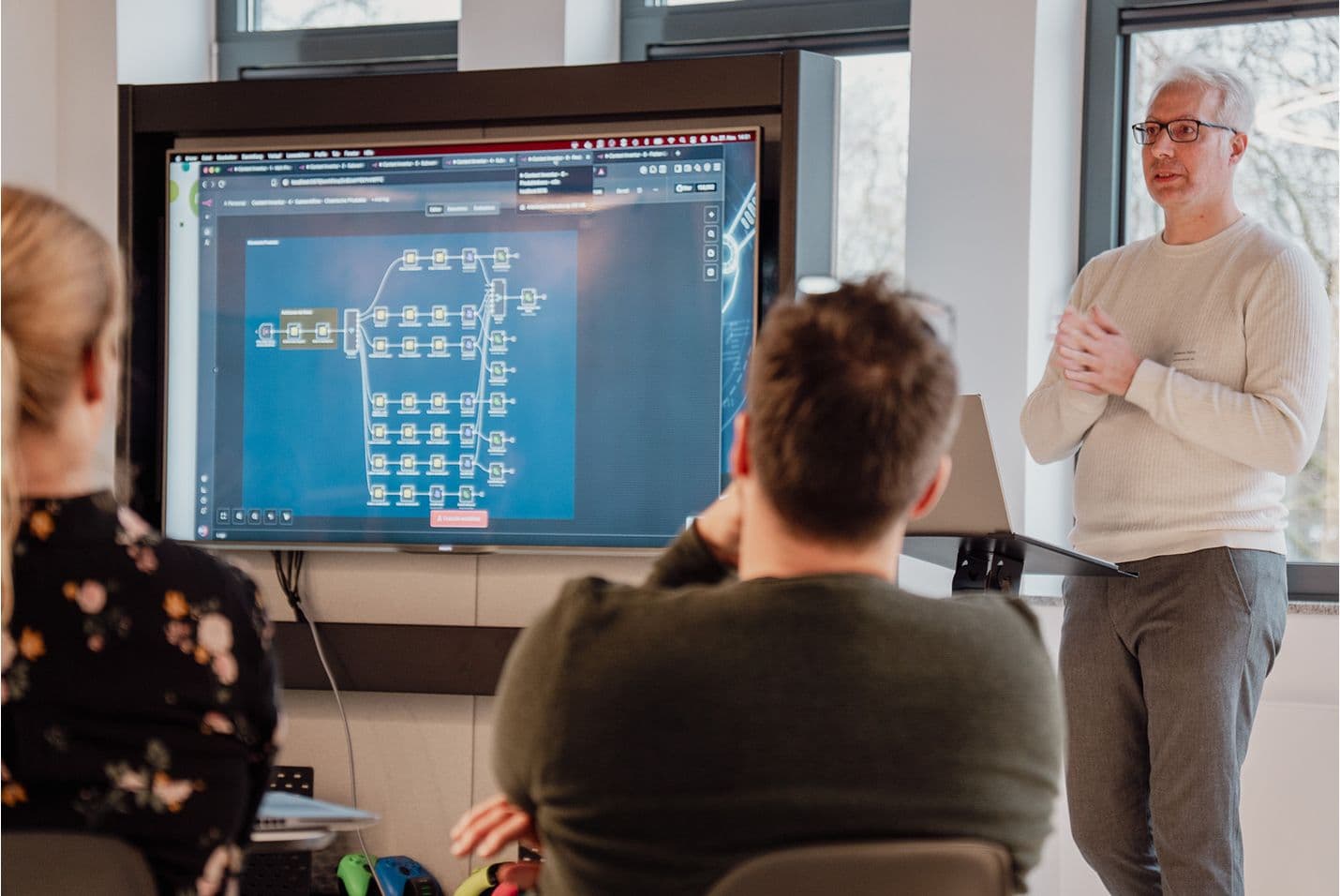 Person gibt eine Präsentation vor zwei sitzenden Teilnehmenden. Auf einem großen Bildschirm ist ein komplexes Diagramm oder Workflow mit vielen verbundenen Knoten zu sehen.