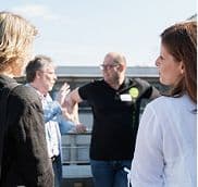 Gruppe von Personen steht draußen zusammen und unterhält sich. Eine Person gestikuliert, während die anderen zuhören. Im Hintergrund sind Gebäudestrukturen und ein klarer Himmel zu sehen.