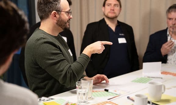 Mehrere Personen sitzen und stehen an einem Tisch während eines Workshops. Eine Person zeigt gestikulierend auf etwas auf dem Tisch, vor ihr liegen Gläser, Stifte, Notizen und farbige Punkte‑Sticker.