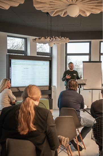 Person steht vorne in einem modernen Seminarraum und präsentiert Inhalte auf einem großen Bildschirm, während mehrere Teilnehmende auf Stühlen sitzend zuhören. Neben dem Bildschirm steht ein leeres Flipchart, und der Raum ist hell mit großen Fenstern und dekorativer Beleuchtung.